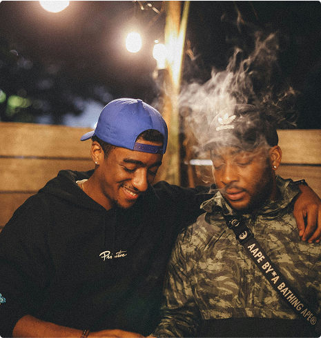 Two young men sitting closely together outdoors at night, one wearing a blue cap and smiling, while the other is exhaling smoke