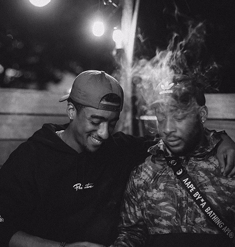 Two young men sitting closely together outdoors at night, one wearing a blue cap and smiling, while the other is exhaling smoke